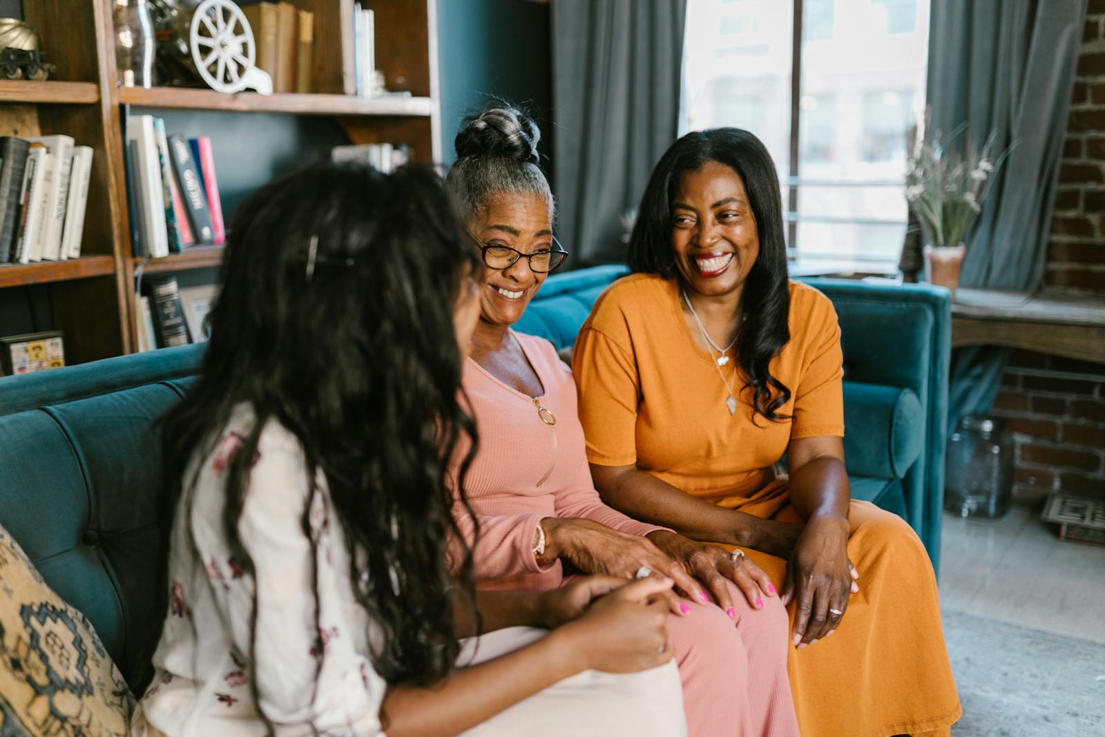 Three women across generations smiling and talking together on a couch in a warm, sunlit home